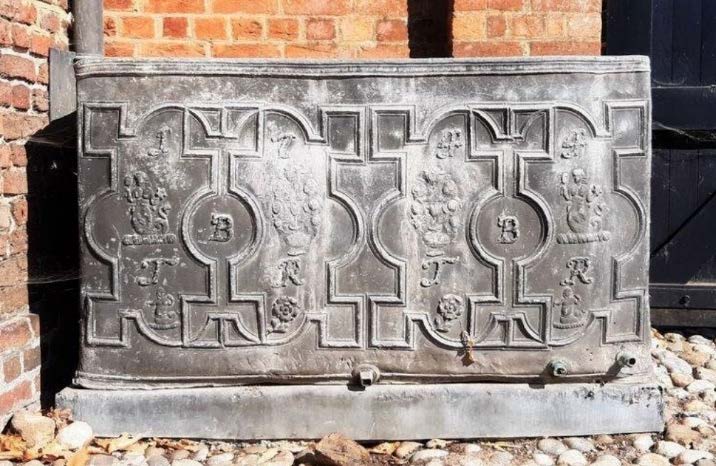A close-up photograph of an ornate cast iron stove or range with relief carvings and crests, resting on gravel near a brick wall.