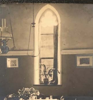 An interior photograph showing a tall, Gothic-style arched window with a plant on the sill, flanked by framed pictures and a hanging lamp.