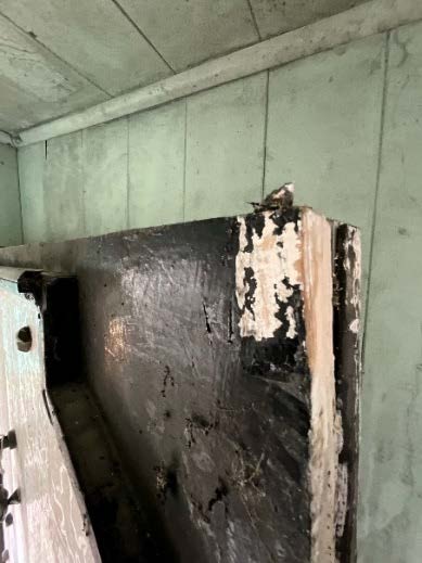 An interior photograph showing a weathered, dark wooden structure with peeling paint and a small bird perched on top, set against pale green paneled walls.