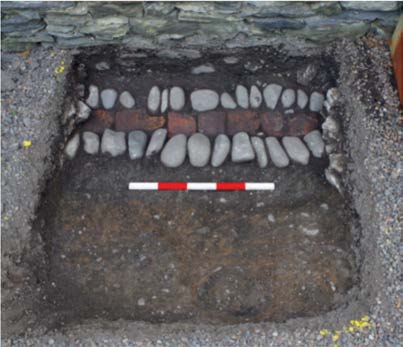 A photograph showing a square excavation pit revealing a stone and brick foundation structure with a red and white scale bar.