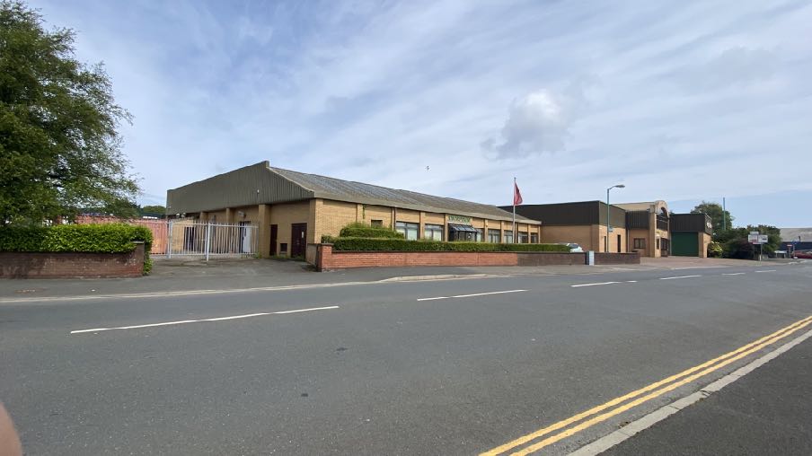 A street-level photograph showing a single-story commercial building with a brick facade and pitched roof, situated along a road with double yellow lines.
