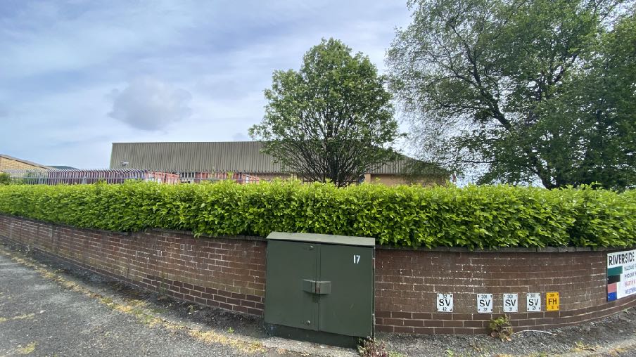 A street-level photograph showing a brick boundary wall topped with a green hedge, with a large industrial-style building visible in the background.