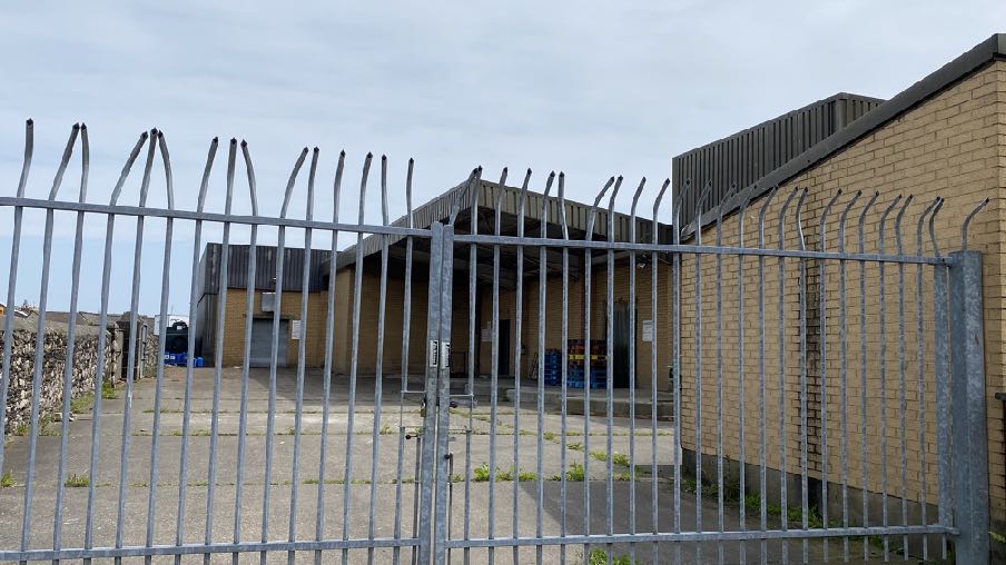 A photograph showing a single-story brick industrial building viewed through a metal security fence with anti-climb spikes.