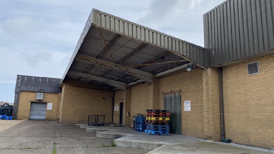 A photograph showing the exterior of a yellow brick industrial building with a large corrugated metal loading bay canopy and stacked pallets.