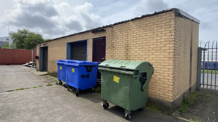 Exterior photograph of a single-story yellow brick building featuring roller shutter doors and large waste bins in a paved yard.