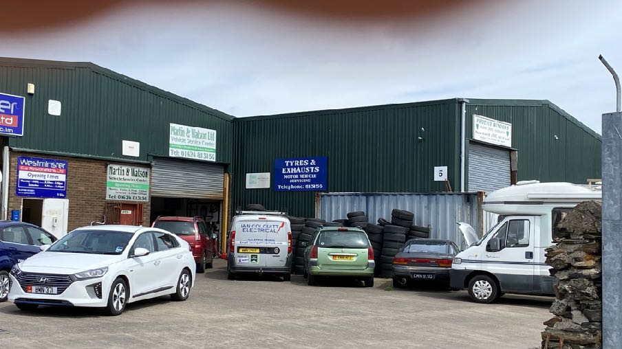 A photograph showing the exterior of a green industrial unit with vehicle service signs and several cars parked in front.