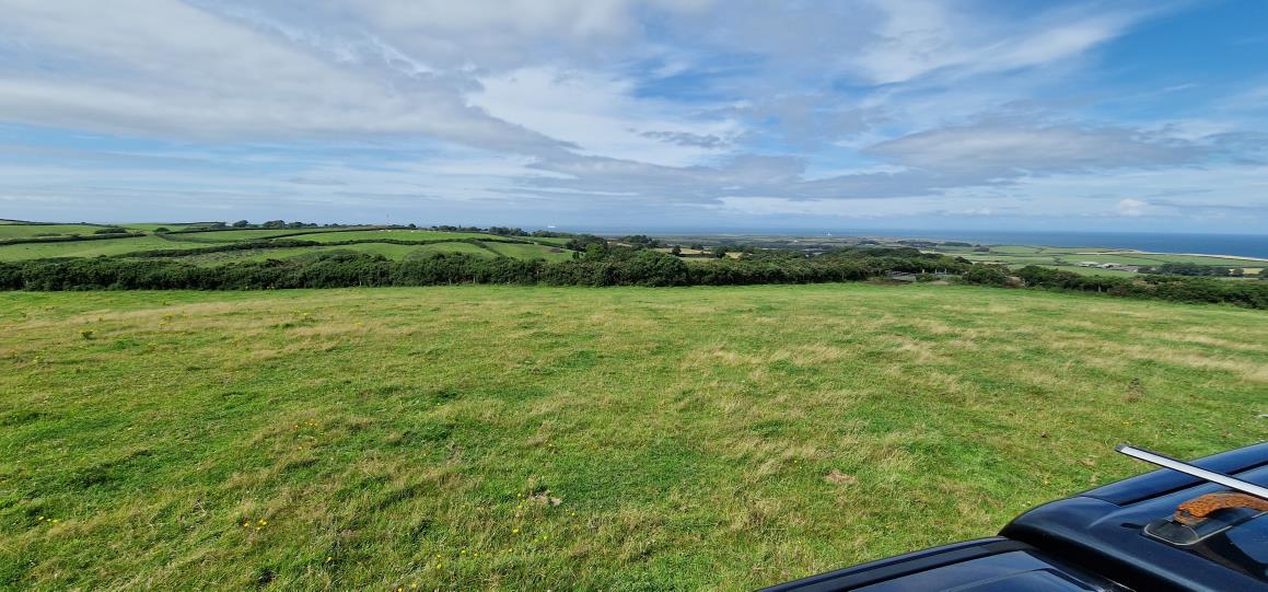 A panoramic photograph of a grassy field and rolling green countryside with hedgerows, looking out towards the sea in the distance.