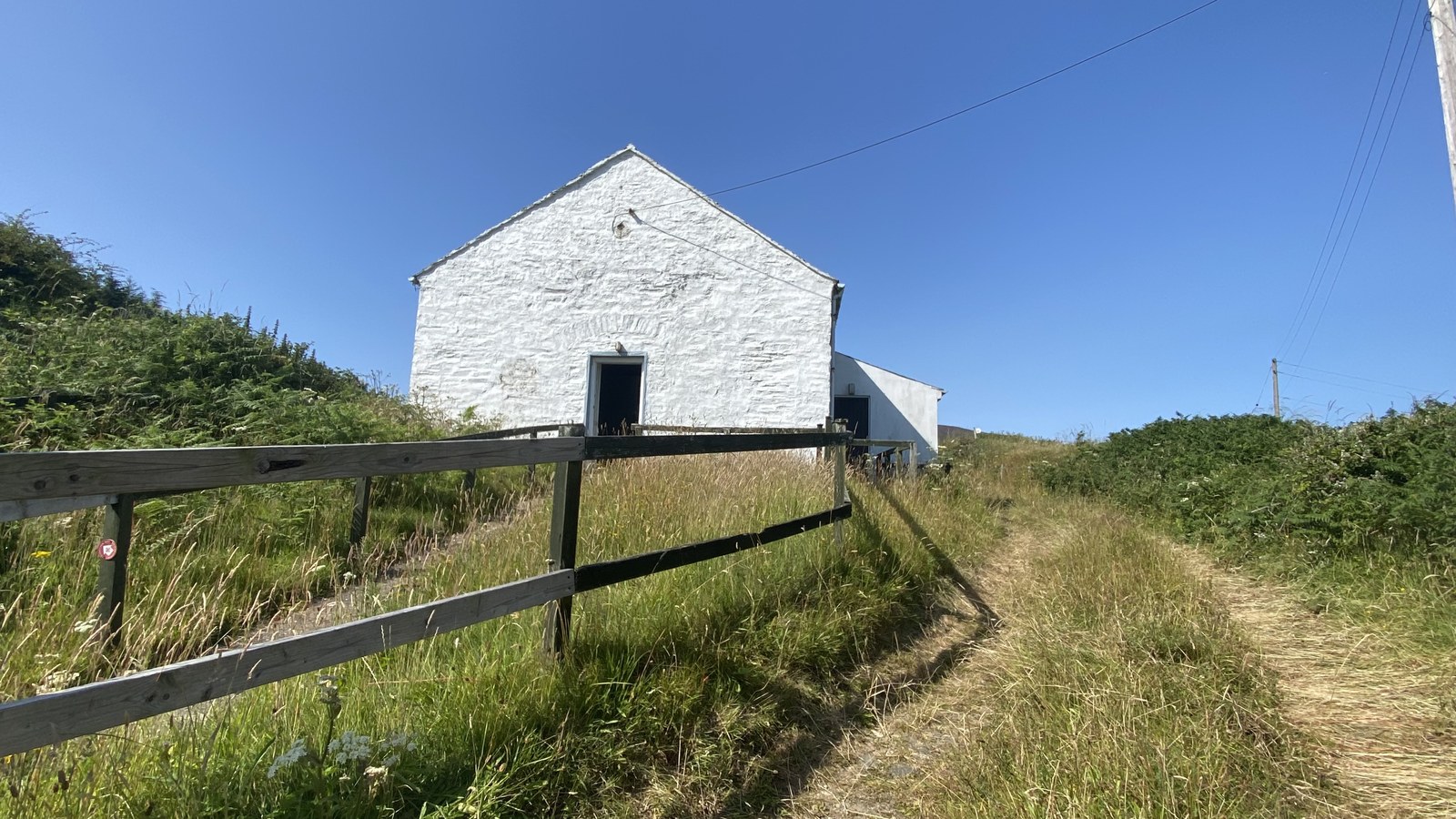A white stone building with a gable roof stands in a grassy rural field, viewed from behind a wooden fence.