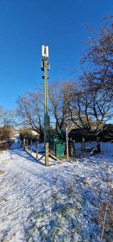 A photograph showing an existing green telecommunications mast with panel antennas and ground equipment cabinets enclosed by a wooden fence in a snowy rural setting.