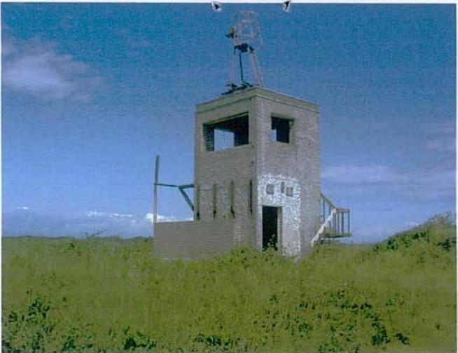 A photograph showing a small, concrete tower-like structure situated in a grassy field under a blue sky.