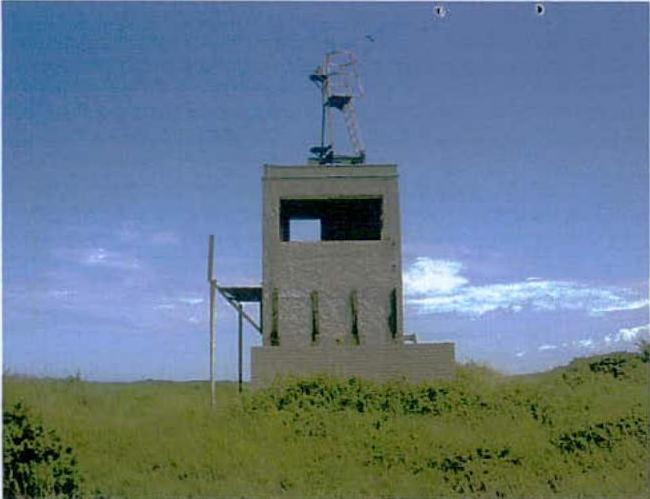 A photograph showing a concrete tower-like structure standing in a grassy field under a blue sky.
