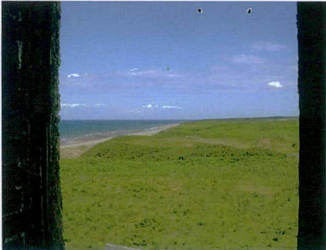 A photograph taken from a dark window frame looking out over a grassy field towards a sandy beach and the sea.