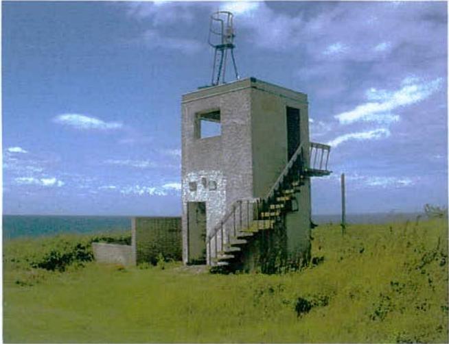 A photograph of a small, concrete coastal tower or military observation post with an external staircase, situated in a grassy field with the sea in the background.