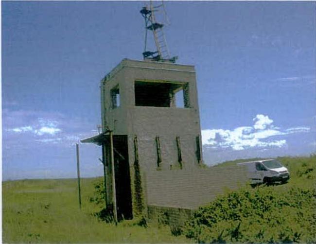 A photograph showing a tall, rectangular concrete tower structure situated in a grassy field with a white van parked nearby.