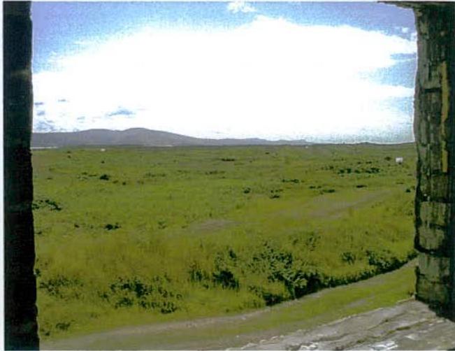 A photograph taken from inside a stone structure looking out over a grassy field towards distant hills.