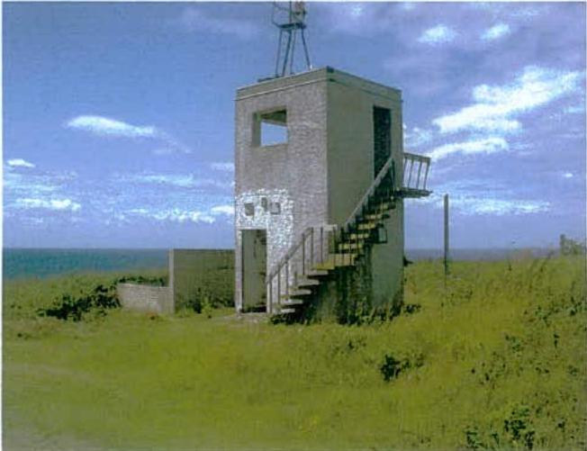 A photograph showing a small, concrete tower-like structure with an external metal staircase, situated in a grassy field with the sea in the background.