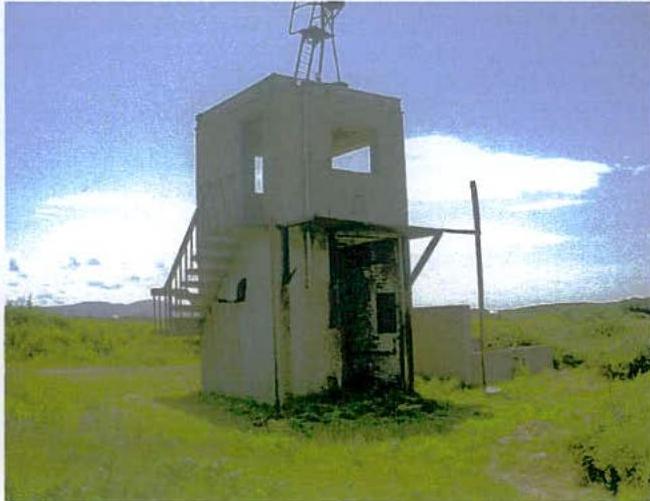 A photograph showing a small, white, concrete tower-like structure standing in a grassy field with an external metal staircase.