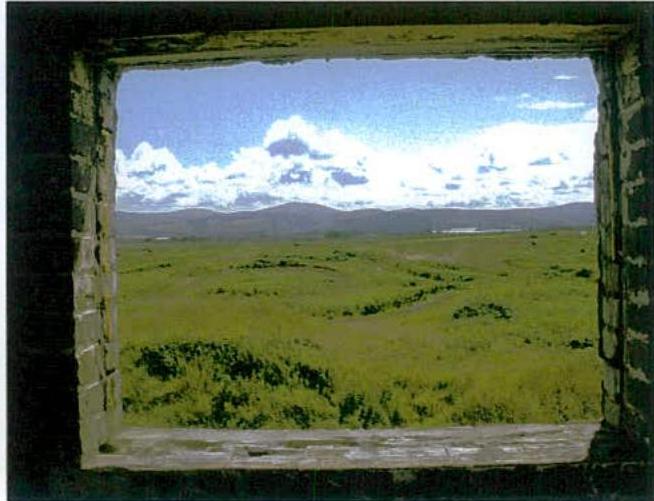 A photograph taken from inside a stone structure looking out through a rough window opening onto a green, hilly landscape.