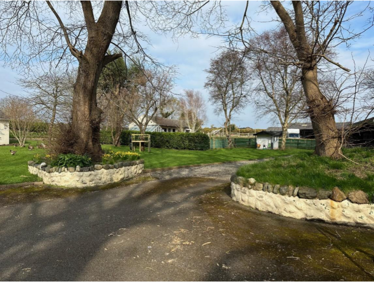 A photograph showing a paved driveway entrance flanked by large trees and low stone walls, leading towards a rural property with existing buildings in the background.