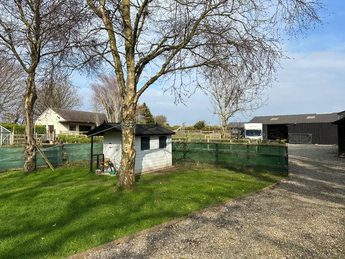 A photograph of a rural property showing a small white outbuilding in the foreground, a detached bungalow to the left, and a large agricultural barn with a truck to the right.