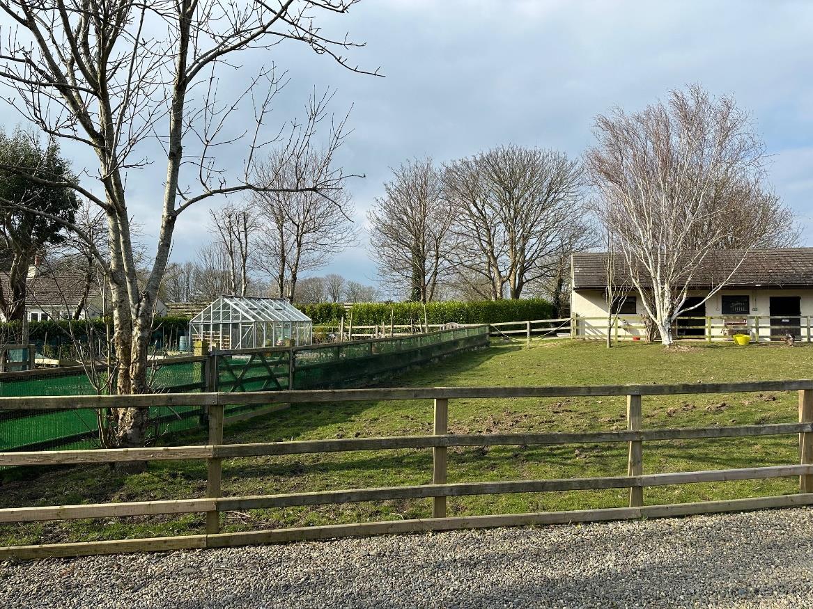 A photograph showing a rural garden scene with a wooden fence, a greenhouse, and a bungalow in the background.
