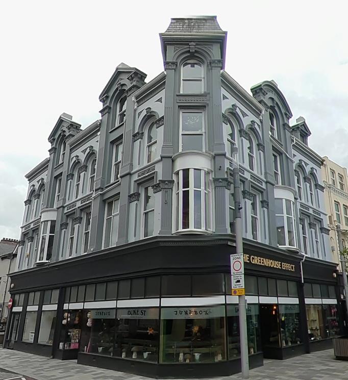 A street-level photograph of a grey corner building with ornate upper floors and ground floor retail units. The building features arched windows and shop fronts, including 'The Greenhouse Effect'.