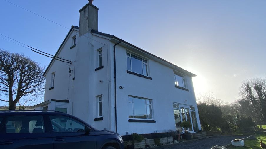 A low-angle photograph of a white, two-story detached house with dark window frames and a blue car parked on a gravel driveway in the foreground.