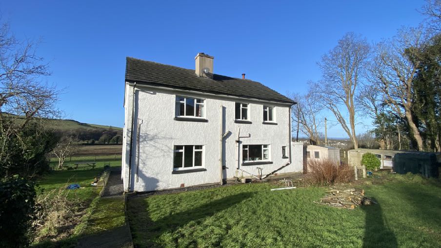 A photograph of a white two-story detached house situated in a rural setting with green fields and trees in the background.