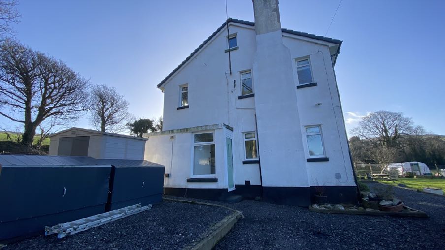 A photograph showing the existing white two-story detached house with a gravel driveway and rural surroundings.