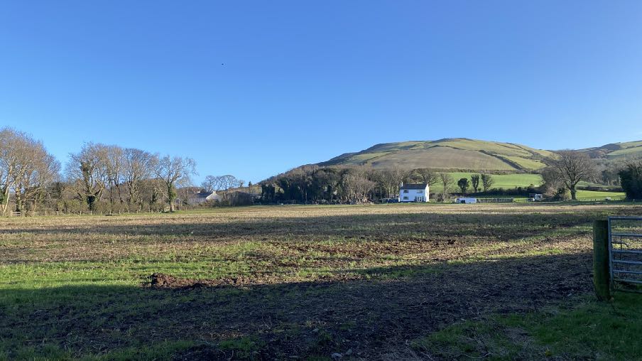 A wide-angle photograph of a rural landscape featuring a white detached house in the distance, surrounded by fields and trees with a large hill in the background.