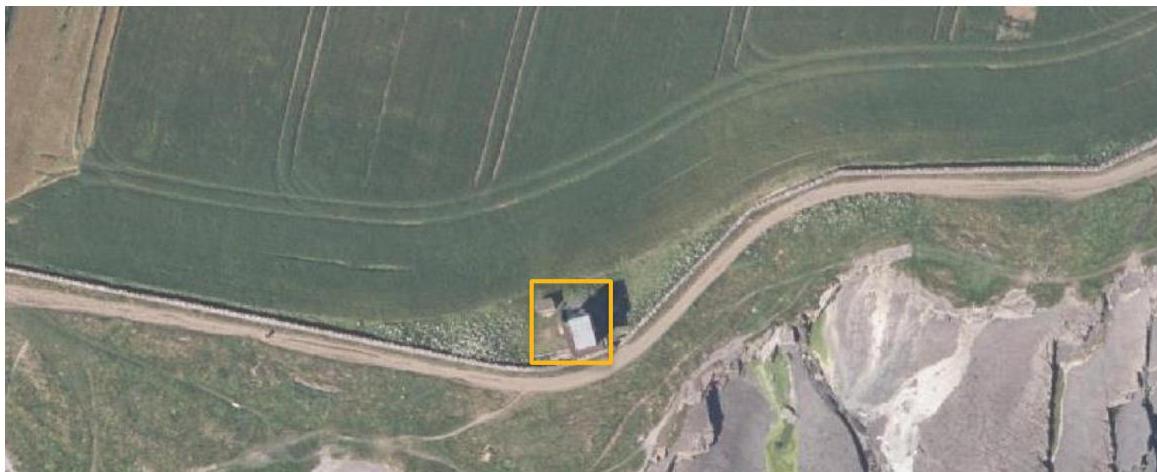 Aerial satellite view of a rural site in Malew, highlighting a small building with a yellow box near a road and rocky terrain.