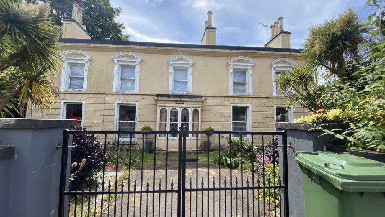 A street-level photograph showing the front elevation of a large, two-story cream-colored detached house behind a black metal gate and grey boundary wall.
