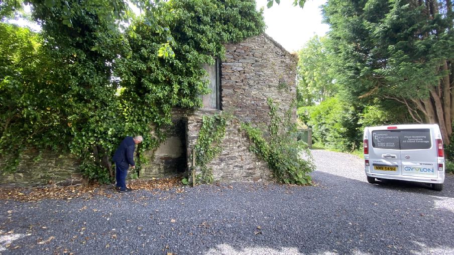 A photograph showing a stone outbuilding covered in ivy with a person inspecting the wall and a work van parked on a gravel driveway.