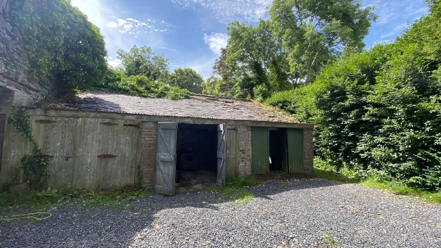 A photograph showing a dilapidated stone and brick outbuilding with wooden doors, situated alongside a gravel driveway and surrounded by dense vegetation.