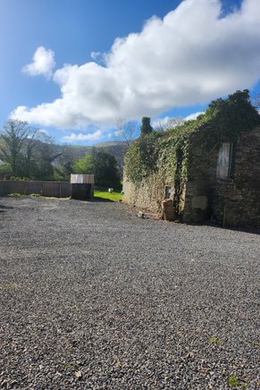 A photograph showing a gravel driveway area adjacent to an old stone building covered in ivy, with a wooden fence and hills in the background.