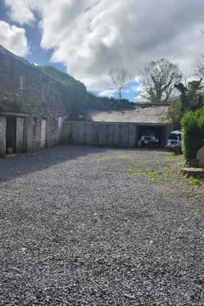 A photograph showing a gravel courtyard or driveway area with stone buildings on the left and rear, set in a rural environment.