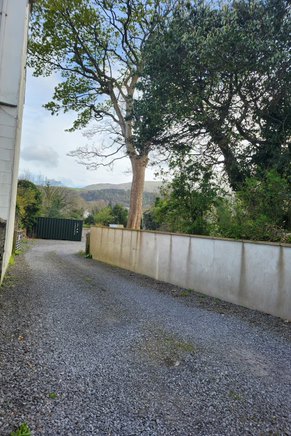 A photograph showing a gravel driveway running alongside a concrete boundary wall with trees and hills in the background.