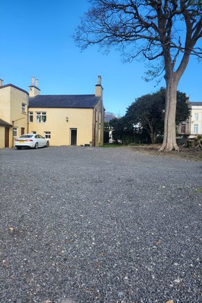 A photograph showing a gravel driveway leading up to a detached yellow house with a dark roof. A white car is parked on the left and a large tree stands on the right side of the frame.