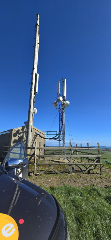 A photograph showing an existing telecommunications mast with panel antennas and a wooden utility pole, situated next to a small building and wooden fence in a rural setting.