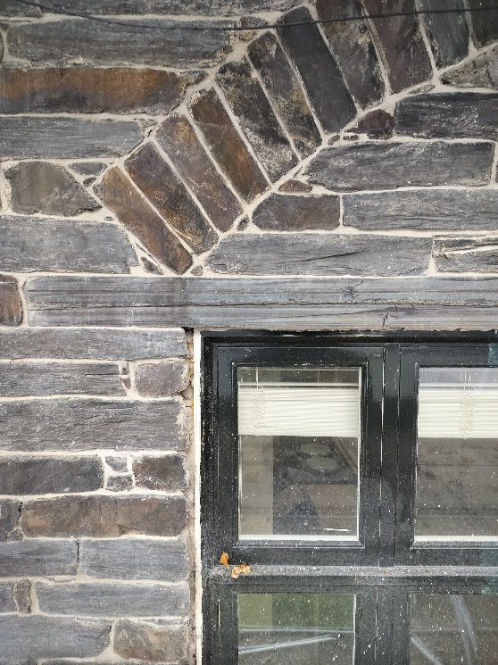 Close-up photograph of a traditional slate stone wall featuring a black-framed window and a wooden lintel.