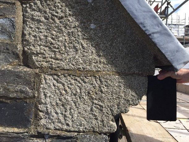 A close-up photograph showing rough-hewn stone masonry on a wall, likely part of a garage or outbuilding, with scaffolding visible in the background.