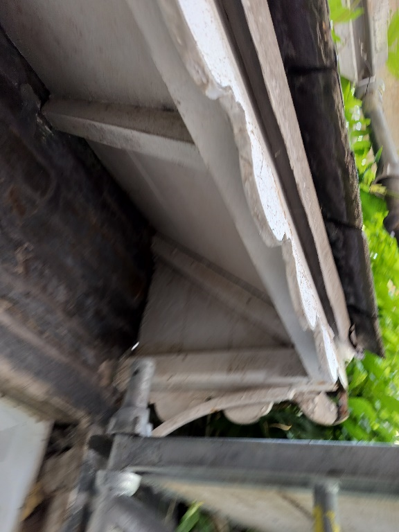 A close-up, angled photograph showing the white painted eaves and decorative wooden brackets of a roof structure, with green foliage visible in the background.