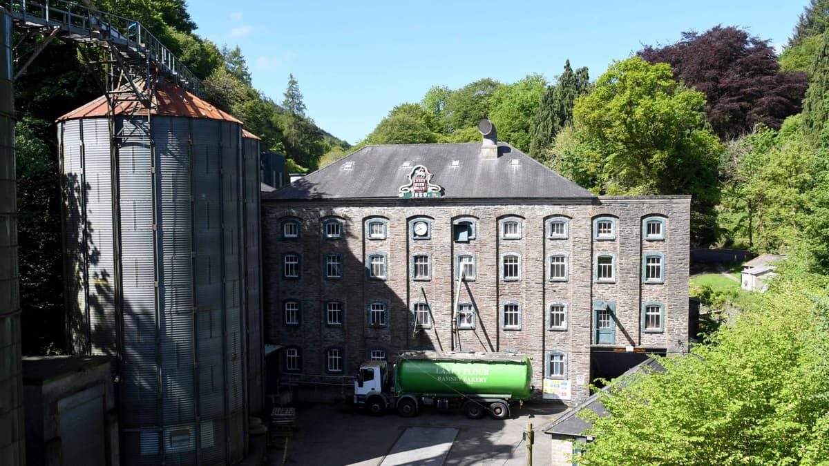 A photograph showing a large, multi-story stone industrial building with a crest on the gable, situated next to a tall metal silo structure with a green truck parked in front.