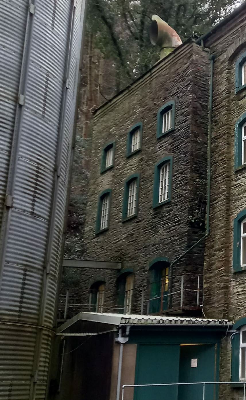 A photograph showing the exterior stone facade of a building with green-framed windows, adjacent to a large corrugated metal structure.