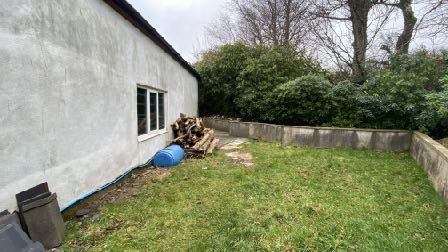 A photograph showing the side elevation of a white rendered building, likely a garage, adjacent to a grassy yard area with a wooden fence and stacked logs.