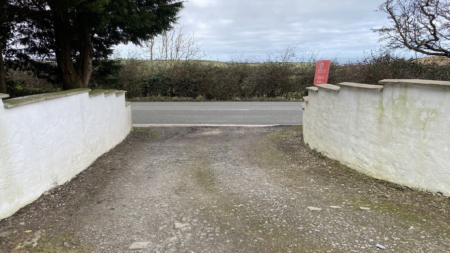 A photograph showing a gravel driveway entrance flanked by white walls leading to a road, with a hedge and field in the background.