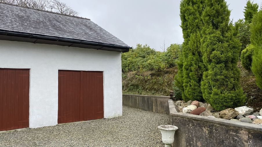 A photograph showing the exterior of a white garage building with two large reddish-brown doors and a slate roof, situated next to a gravel driveway and a stone retaining wall with tall evergreen trees.