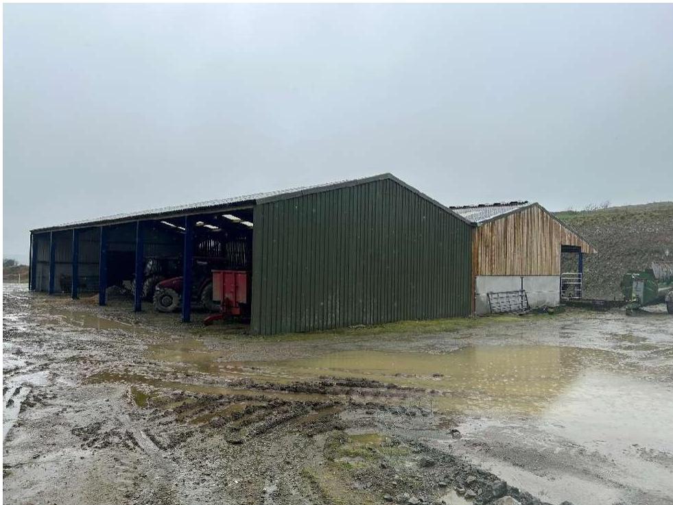 A photograph showing two agricultural buildings situated on a muddy, wet farmyard. The larger structure is a green metal shed with blue supports containing farm machinery, adjacent to a smaller wooden and concrete bui...