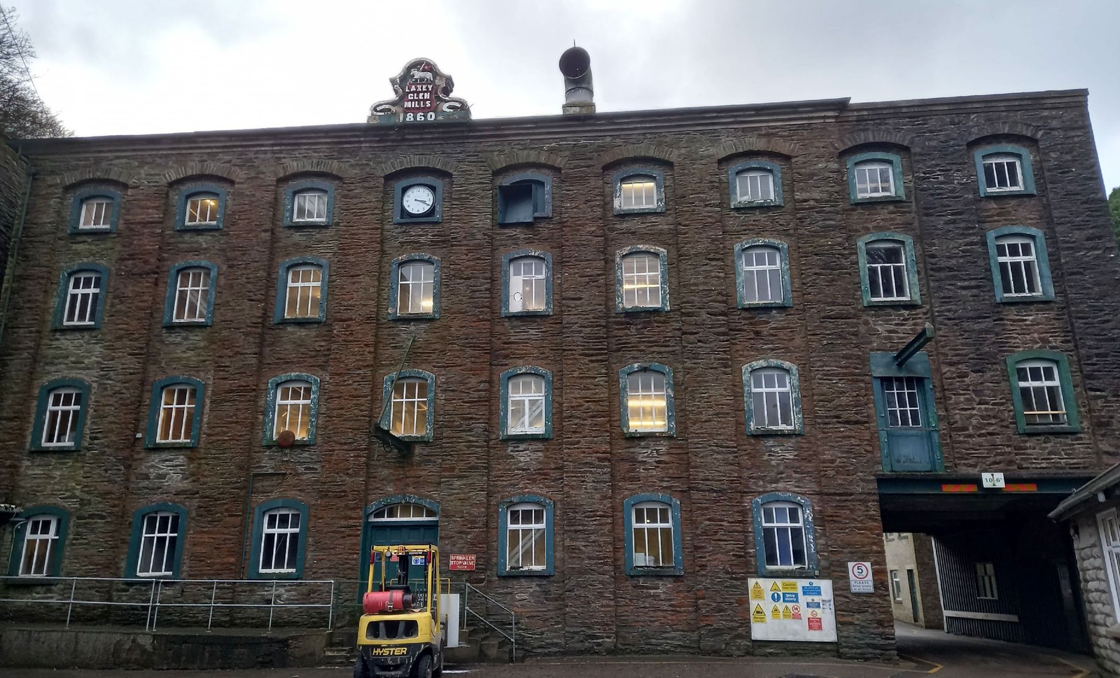 A photograph of the historic Laxey Glen Mills building, a large stone structure featuring a 1860 sign and a clock face on the upper level.