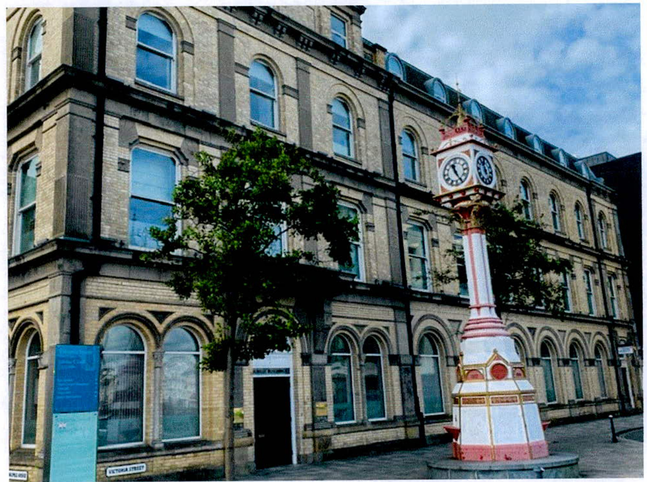A street-level photograph of a large, multi-story stone building featuring arched ground-floor windows and a prominent ornate clock tower in the foreground.